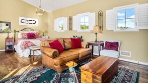 A spacious living room with a leather sofa, a wooden coffee table, and a patterned rug. There are two windows with shutters, a chandelier, and a bookshelf with books. - Distant Views | Incredible Views | Gas Fireplace | Queen Bed (Fredericksburg)