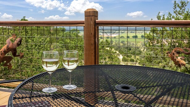 A metal table with two glasses of white wine, a wooden fence, and a scenic view of green fields and trees. - Distant Views | Incredible Views | Gas Fireplace | Queen Bed (Fredericksburg)