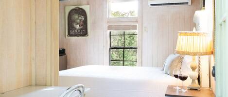 A bedroom with a bed, a window, an air conditioning unit, a lamp, and a small table with glasses.