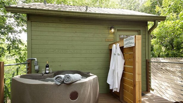 Un jacuzzi con toallas y una botella de vino, una terraza de madera y una pequeña estructura de madera con una puerta y una toalla colgada.