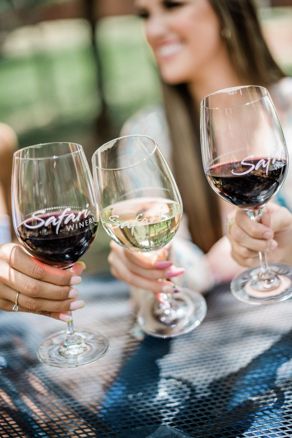 Three wine glasses with 'Safari Wine' branding, held by individuals at a table with a metal mesh surface.