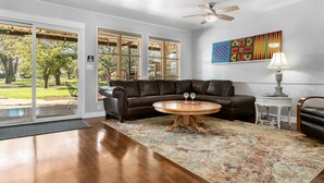 A spacious living room with a leather sofa, a round wooden coffee table, and a large window offering a view of a park. - Harrys House | 3/2 Cottage | Full Kitchen | Hot Tub | In Town (Fredericksburg)