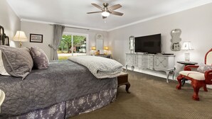A bedroom with a bed, a ceiling fan, a TV, a dresser, a chair, and a window with a view of greenery. - Harrys House | 3/2 Cottage | Full Kitchen | Hot Tub | In Town (Fredericksburg)