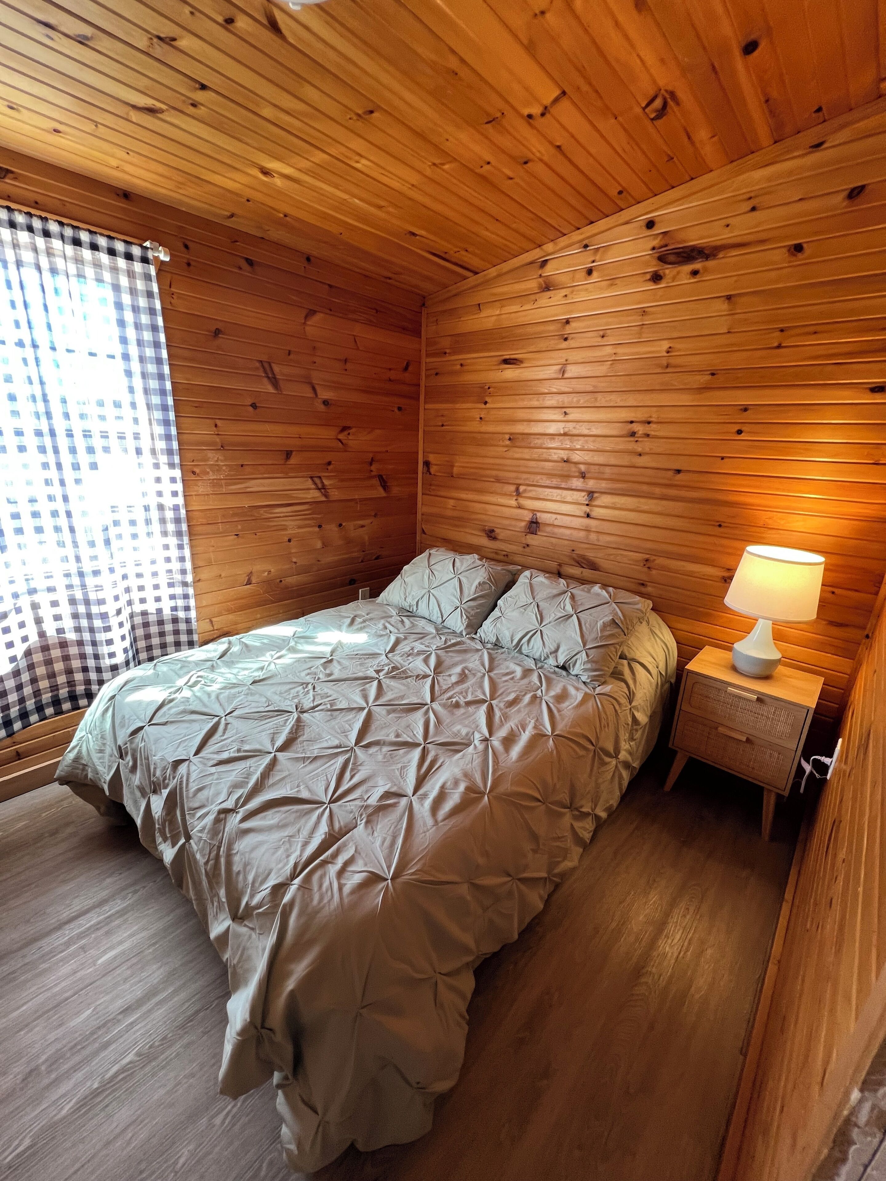 Une pièce de cabane en bois comprenant un lit, une table de chevet et une fenêtre avec des rideaux à carreaux.