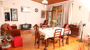 A dining area with a table set for four, surrounded by wooden chairs. A red ottoman and a brown sofa are visible in the foreground. - Oudtshoorn Homestay Accommodation (Oudtshoorn)