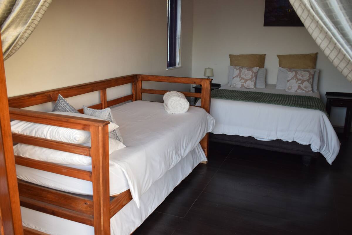 Uma cama de casal e uma cama simples, ambas com cabeceiras de madeira, num quarto com soalho escuro.