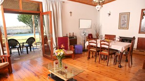 A spacious living room with a glass coffee table, wooden dining table, and chairs. There is a fireplace, a large mirror, and a cross on the wall. - Oudtshoorn Homestay Accommodation (Oudtshoorn)