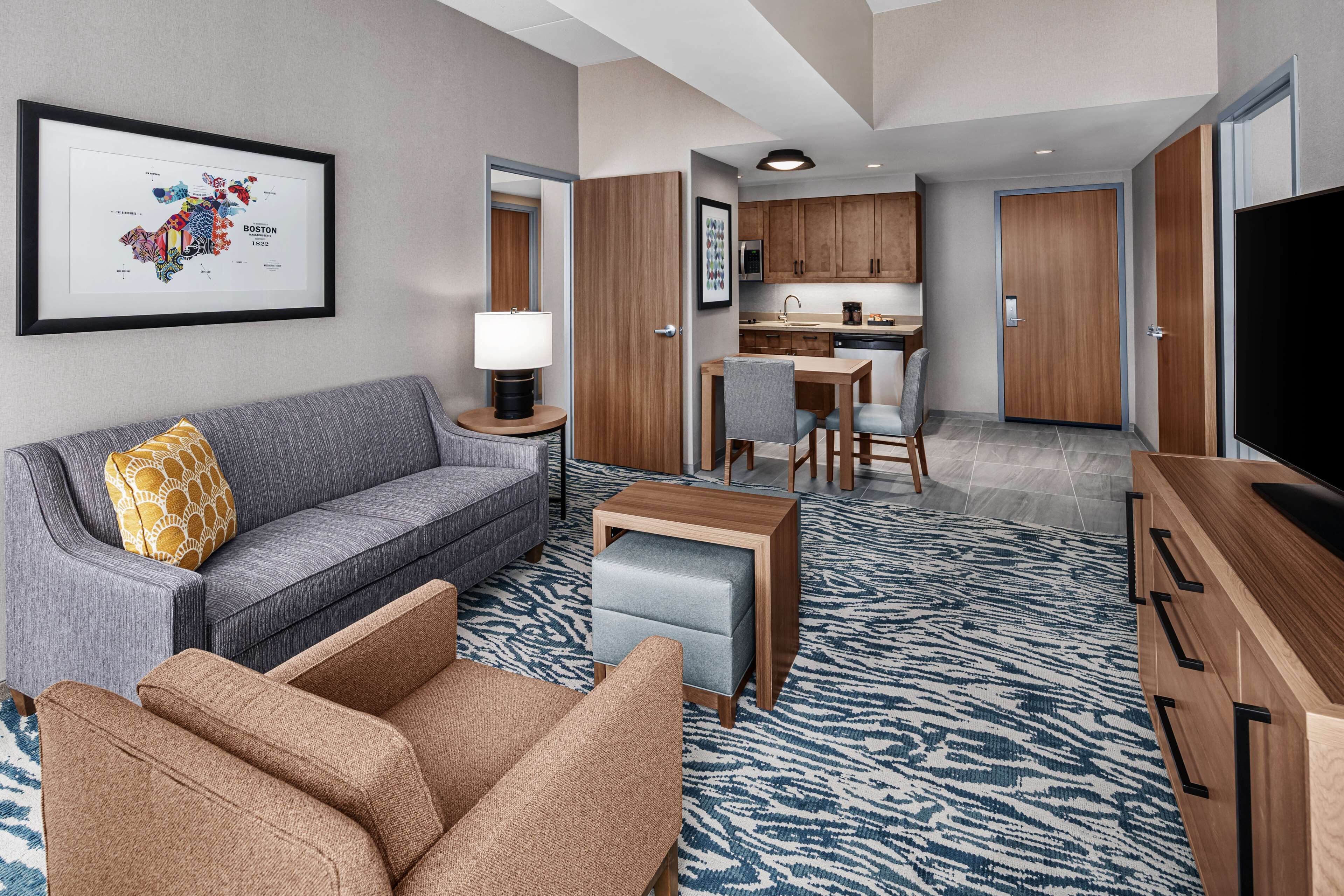 A modern hotel room with a grey sofa, a wooden coffee table, a patterned rug, and a television.