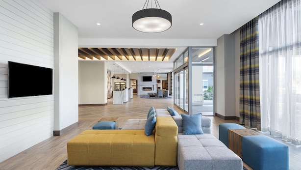 A modern hotel lobby with a large sofa, ottomans, a fireplace, and a TV mounted on the wall. - Homewood Suites by Hilton Boston Woburn (Woburn)