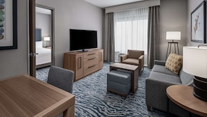 A hotel room with a flat-screen TV, a wooden desk, a sofa, a chair, and a small ottoman. - Homewood Suites by Hilton Boston Woburn (Woburn)