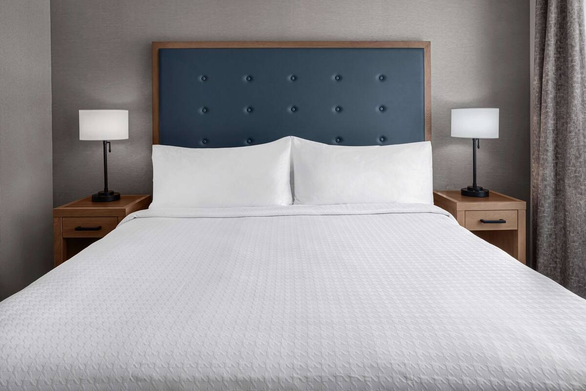 Una cama bien hecha con cabecera capitonada, dos mesitas de noche con lámparas y una mesita auxiliar de madera.