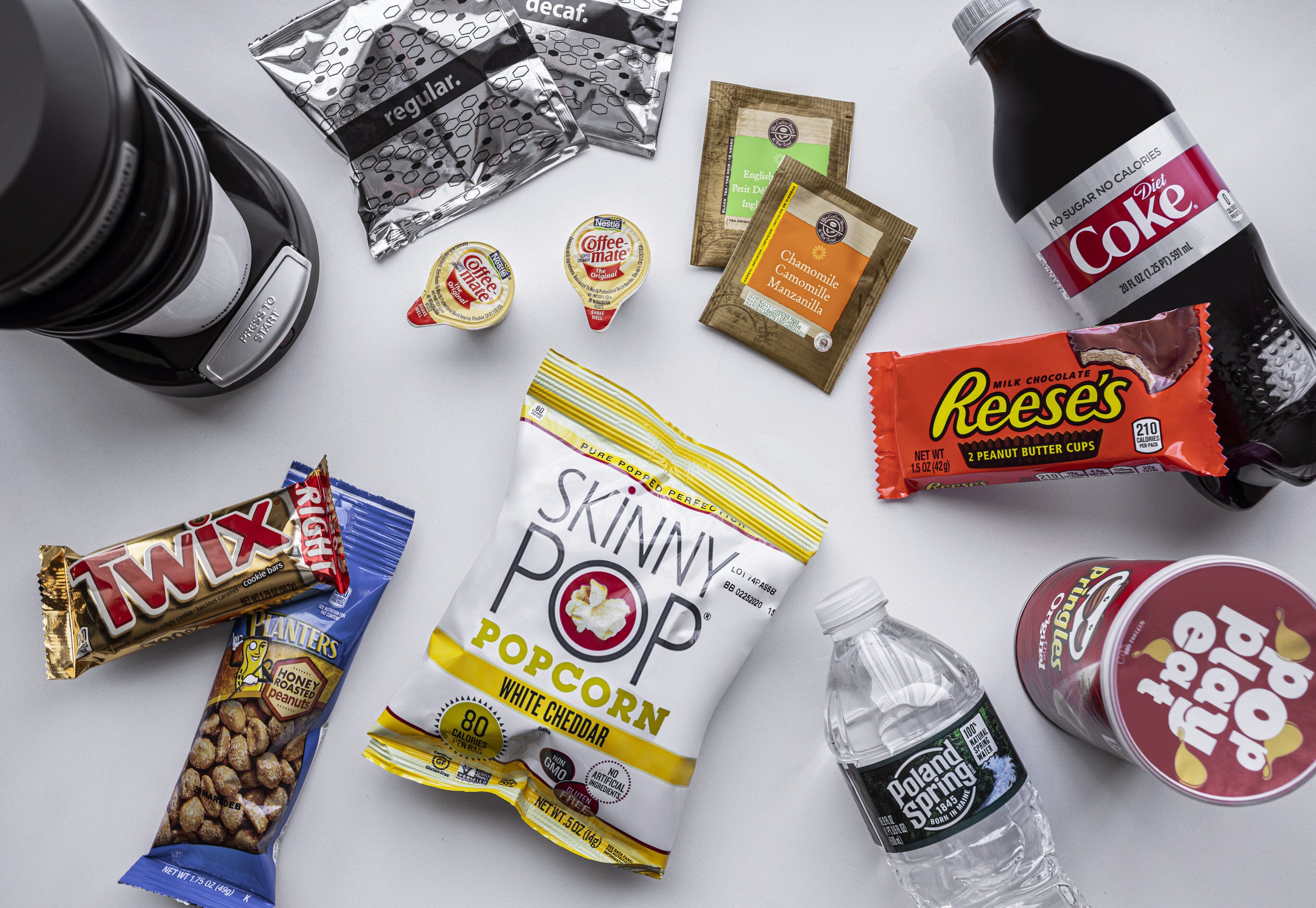 Différentes sortes de collations et de boissons, notamment des barres Twix, du pop-corn Skinny Pop et du Coca-Cola, disposées sur une surface blanche.