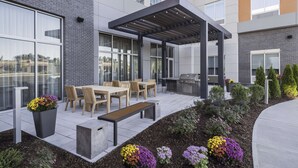 A modern outdoor patio with a wooden table, chairs, a grill, and a pergola. - Homewood Suites by Hilton Boston Woburn (Woburn)