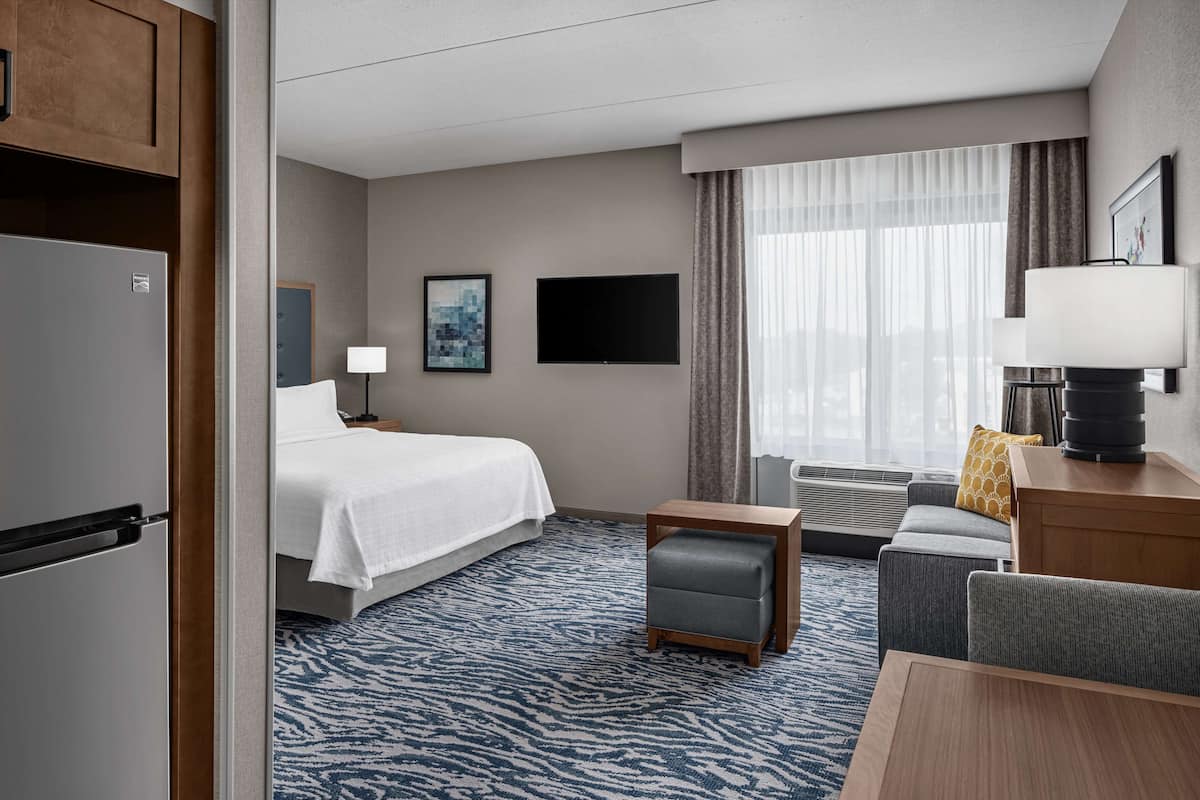 Habitación de hotel con cama, escritorio, silla, una mesita, refrigerador, televisor y ventana con cortinas.