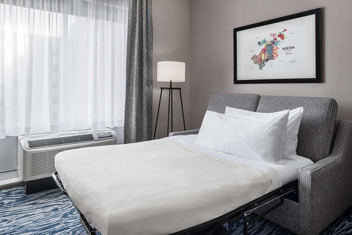 Una habitación de hotel con una cama, una lámpara de pie, un mapa enmarcado de Boston y una ventana con cortinas.