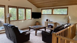 A living room with a wooden ceiling, large windows, a television, and leather sofas. - Cozy Sunriver Home w/ AC, Fireplace, HotTub, 8 SHARC Passes, Outdoor Heaven (Sunriver)