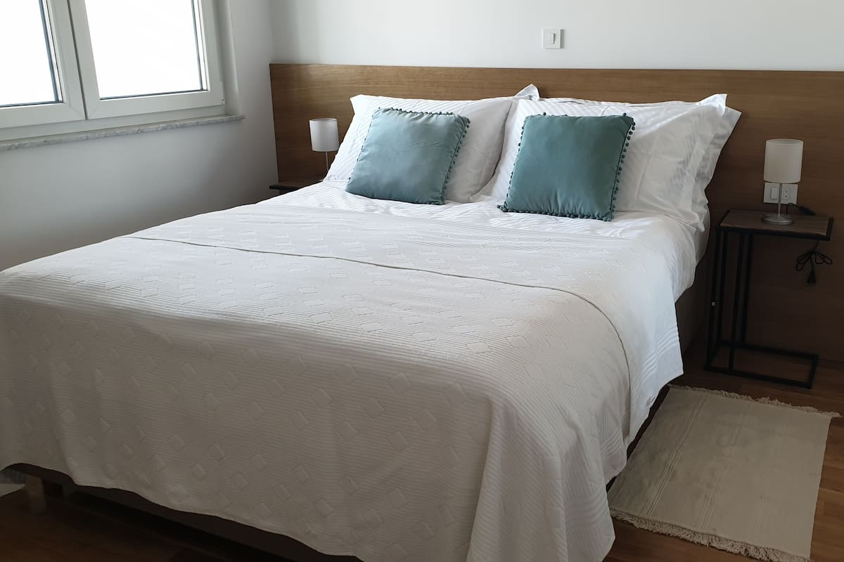 A neatly made bed with a white duvet and two teal pillows, a bedside table with a lamp, and a small rug on the wooden floor.