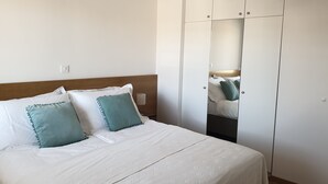 Un lit bien fait, avec une literie blanche et des coussins turquoise, une tête de lit en bois et une armoire blanche dotée d’un miroir.
