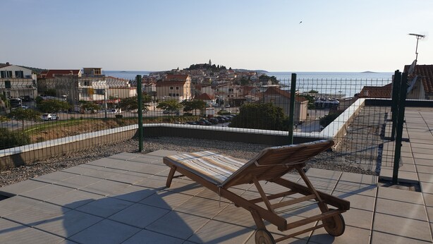Un banc en bois sur une terrasse carrelée, donnant sur une ville côtière avec ses bâtiments et un ciel dégagé.