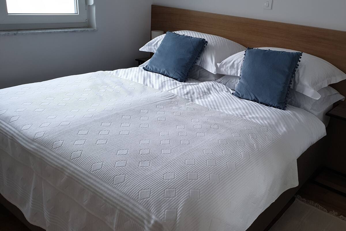 A neatly made bed with white bedding and blue pillows, a bedside table with a lamp, and a window with blinds.