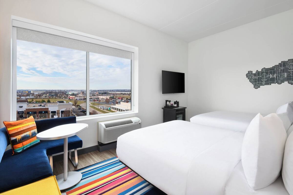 A hotel room with a large window, a bed, a blue sofa, a small table, and a flat-screen TV.