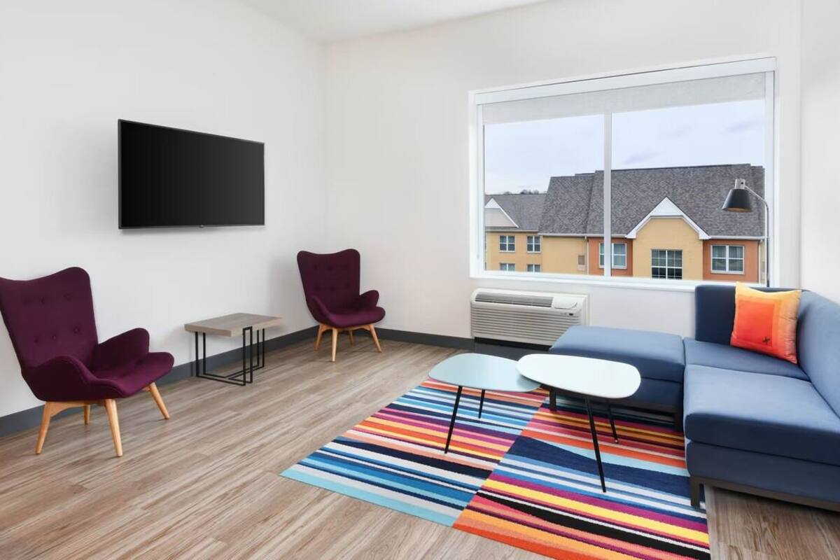 A modern living room with a blue sofa, two purple armchairs, a flat-screen TV mounted on the wall, and a large window with a view of buildings.