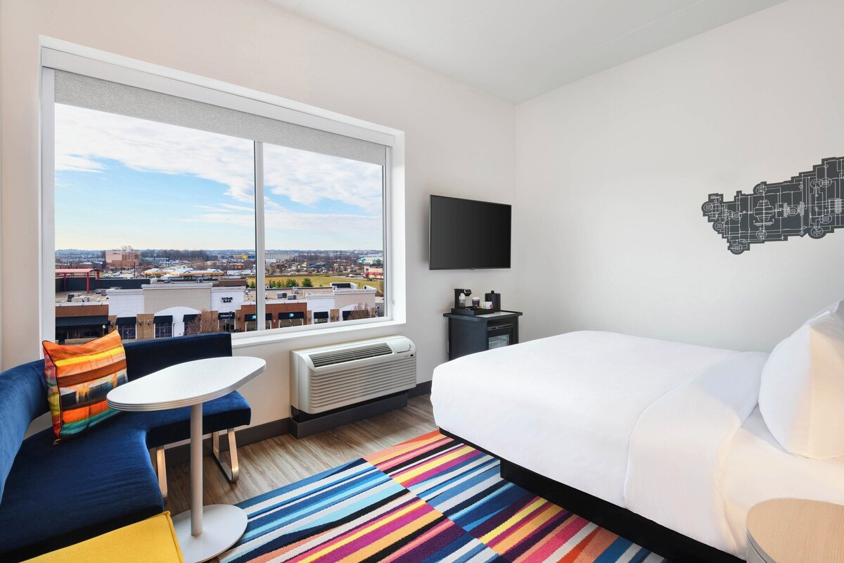 A hotel room with a large window, a bed, a small table, a sofa, and a TV on the wall.