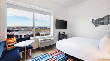 A hotel room with a large window, a bed, a small table, a sofa, and a TV on the wall.