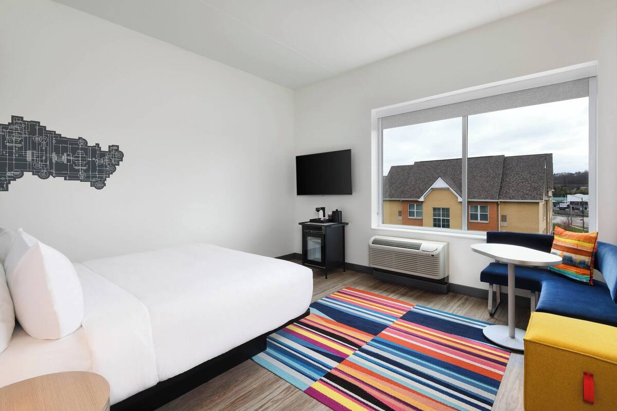 A modern hotel room with a bed, a small table, a sofa, a colorful striped rug, a flat-screen TV, and a window with a view of buildings.