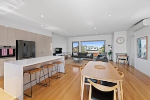 A modern kitchen with a central island, bar stools, a dining table with chairs, a sofa, and a large window.