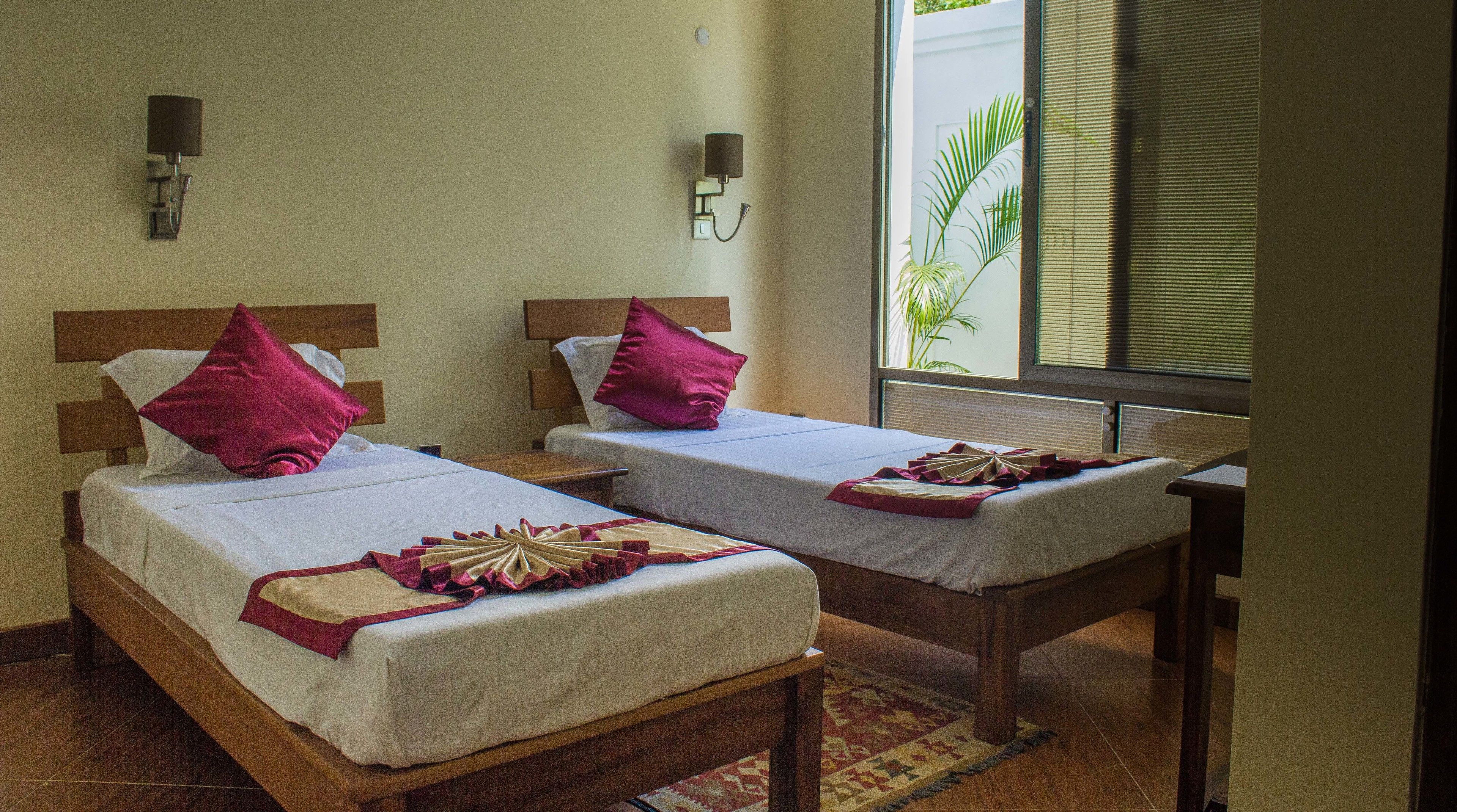 Two single beds with wooden headboards, white linens, and red pillows. There are wall-mounted lamps and a window with blinds.