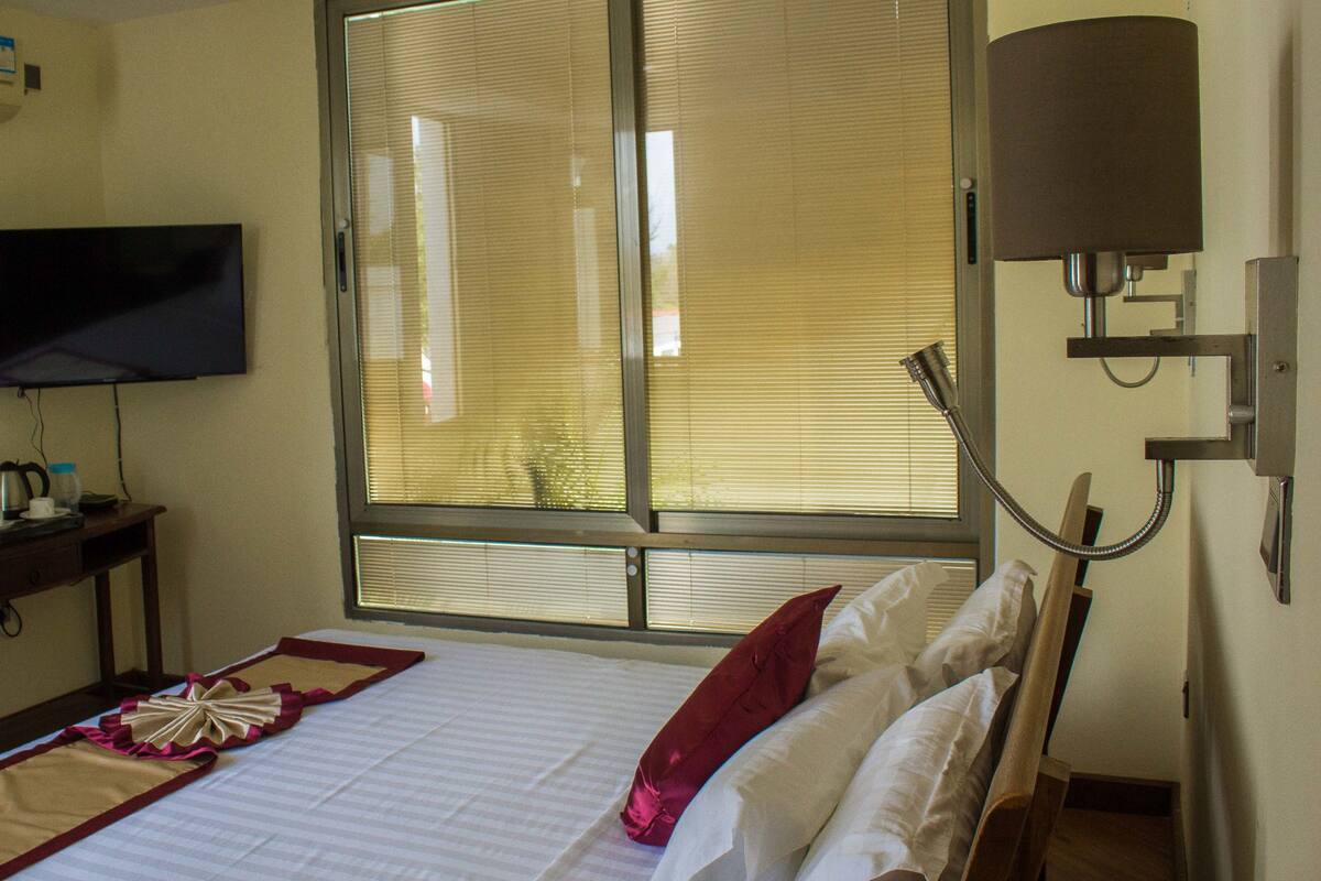Una camera d'albergo con un letto, una televisione, una finestra con persiane e una lampada da comodino.