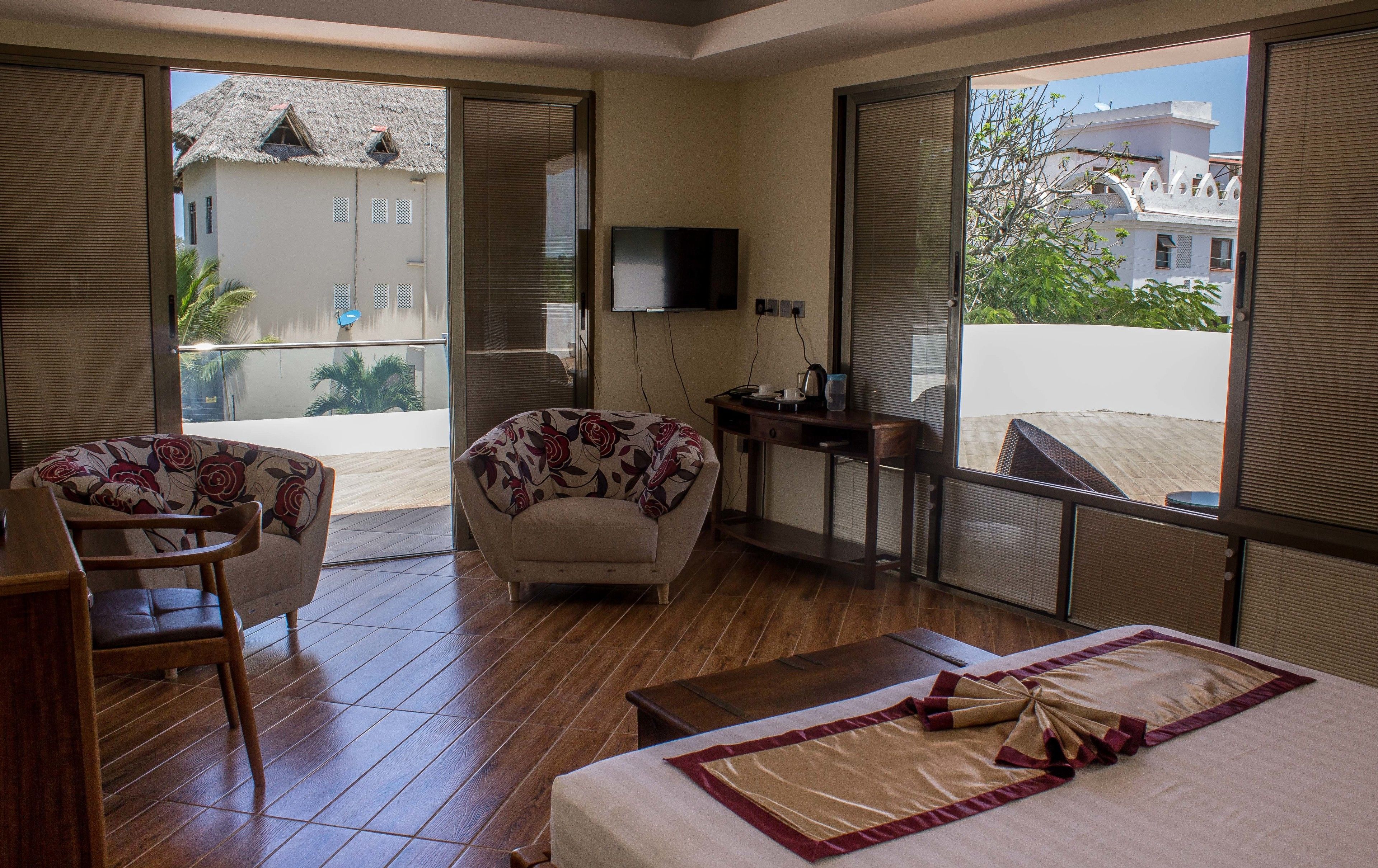 Habitación de hotel con cama, sofá, sillón, una mesita y televisión. Tiene un balcón con vistas a edificios y árboles.