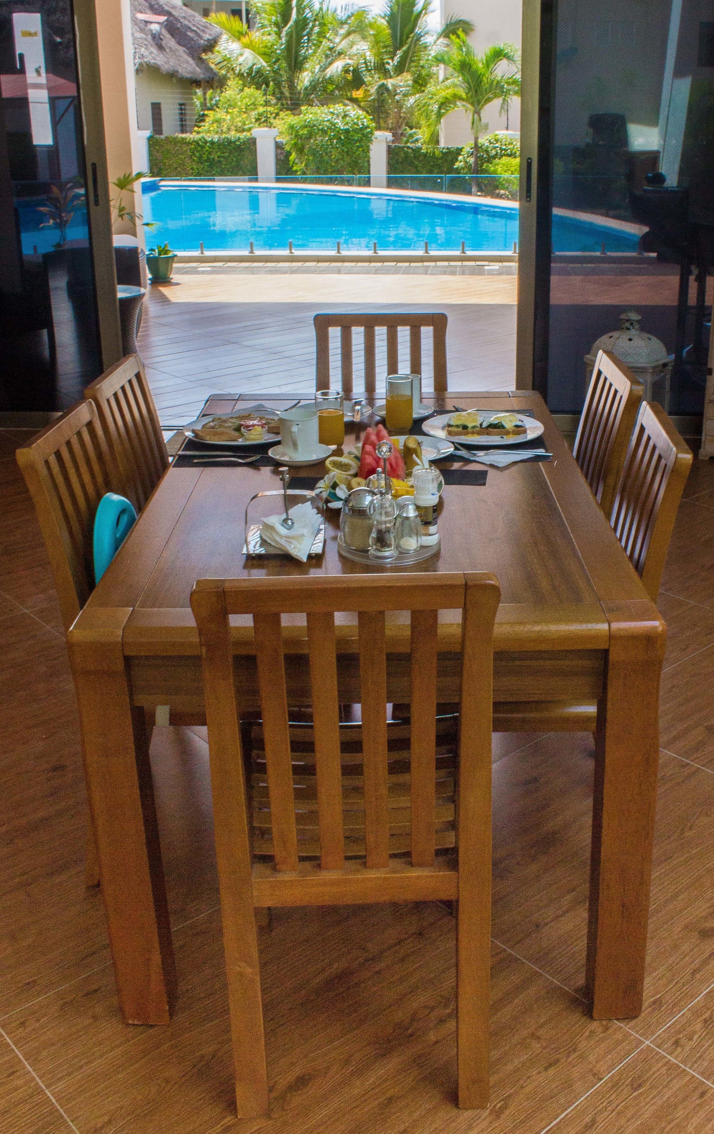 A dining table with chairs, a vase with flowers, and a view of a swimming pool and greenery.