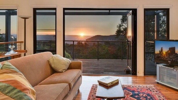A modern living room with a sofa, a coffee table, and a view of a sunset through large sliding doors. - Narrow Neck Views - Spacious Mountains Home with Stunning Views! (Katoomba)