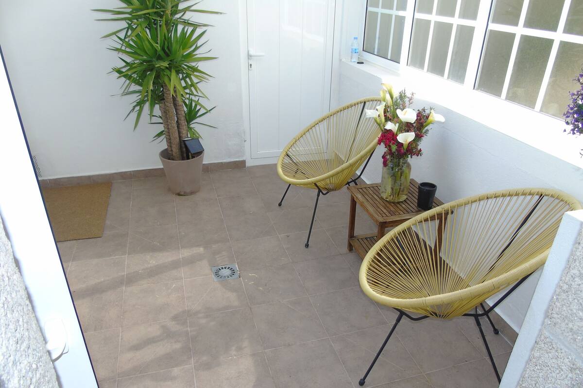 A small patio area with two wicker chairs, a potted plant, and a small table with a vase of flowers.