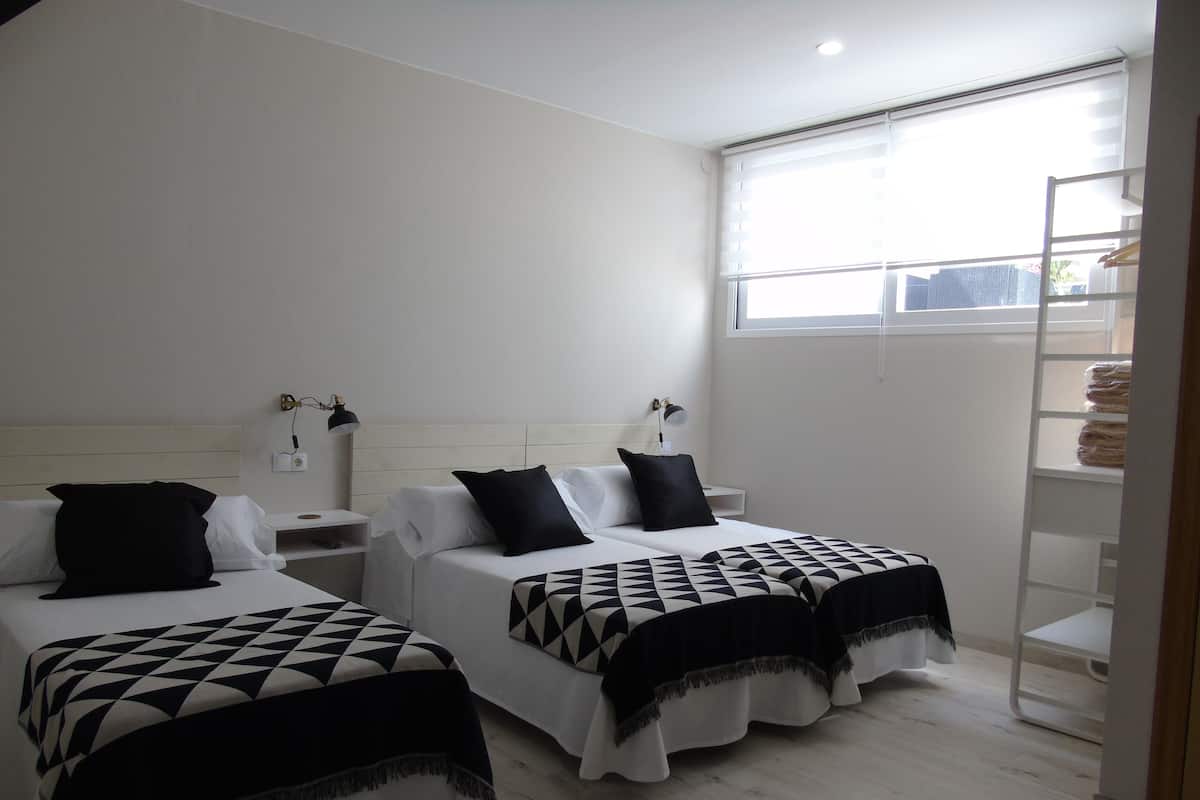 A hotel room with two beds, each with a geometric-patterned bedspread, white bedding, and black pillows. There are bedside tables with lamps, a window with blinds, and a shelving unit against the wall.