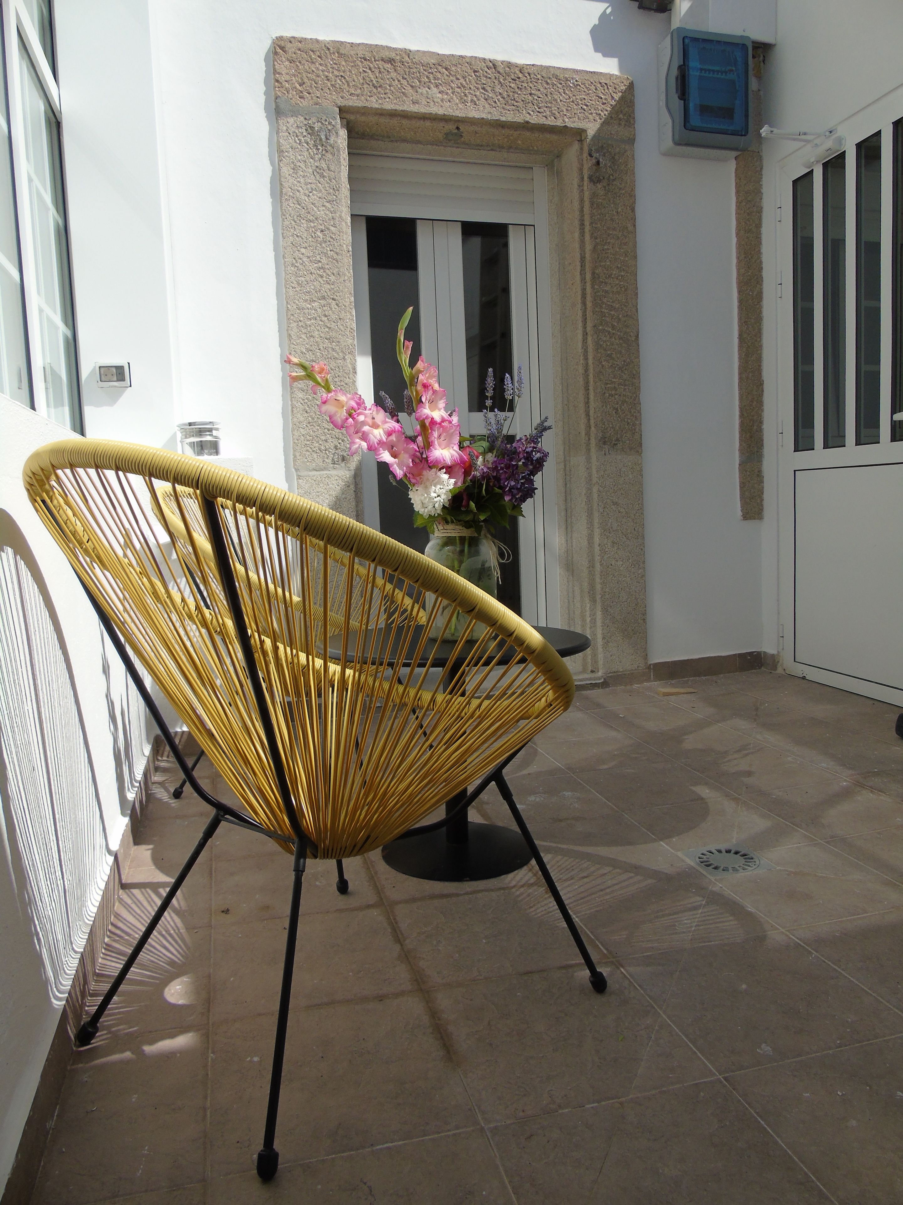 Una silla de mimbre amarilla en un patio empedrado con una puerta blanca y un jarrón de flores al fondo.