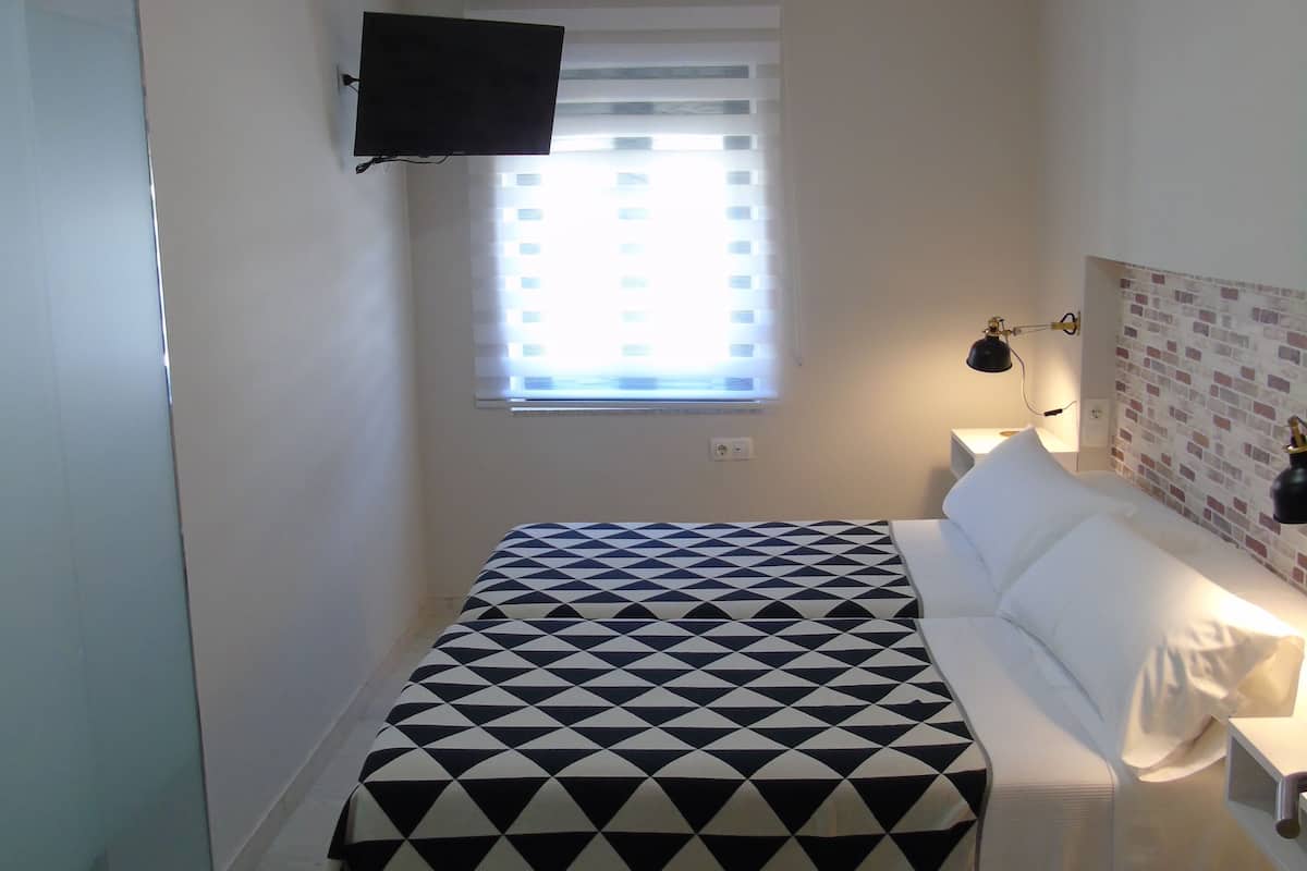 A bedroom with a bed, a TV mounted on the wall, a window with blinds, and a nightstand with a lamp.