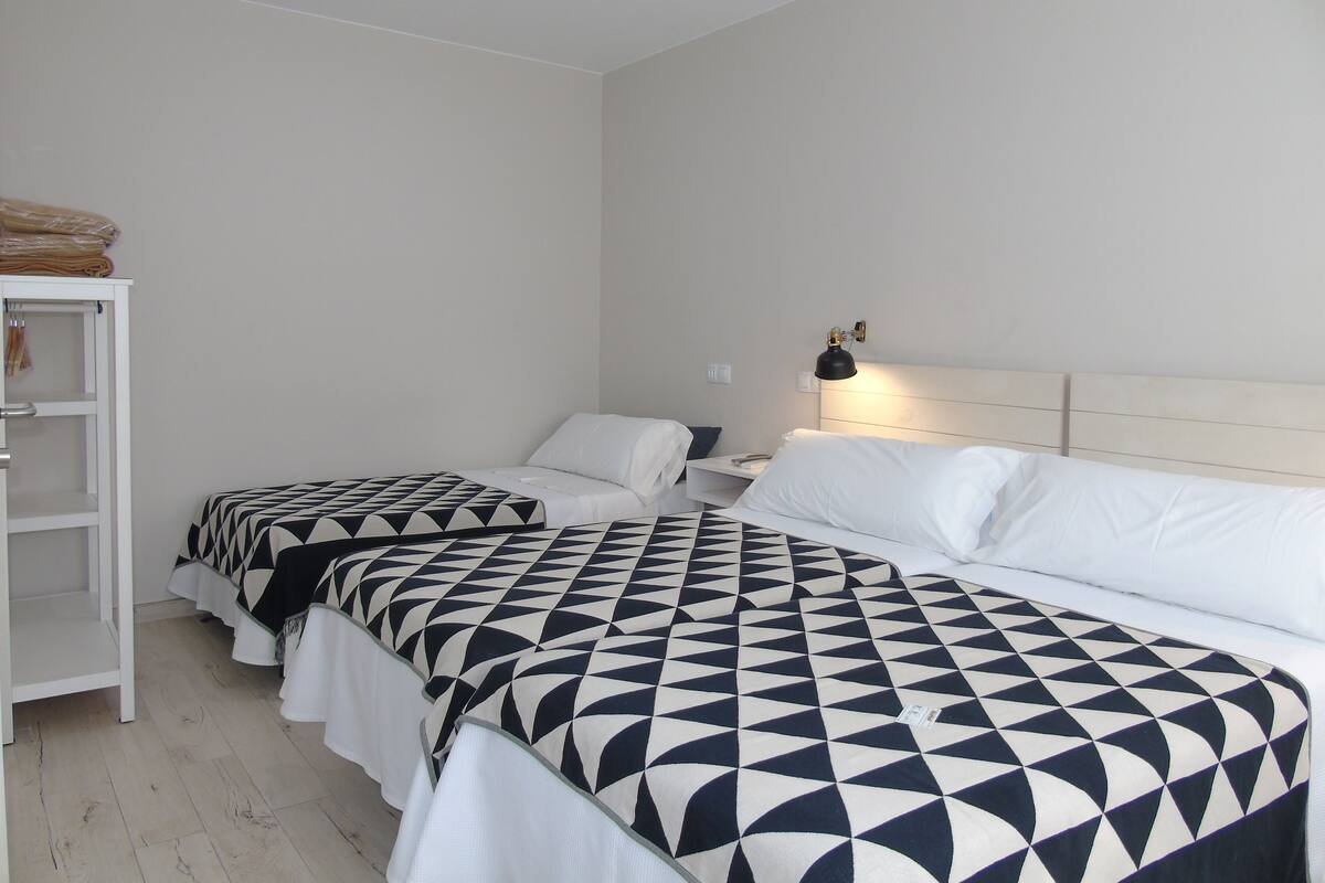 A hotel room with two beds, each with a geometric-patterned bedspread, a bedside table with a lamp, and a white wardrobe.