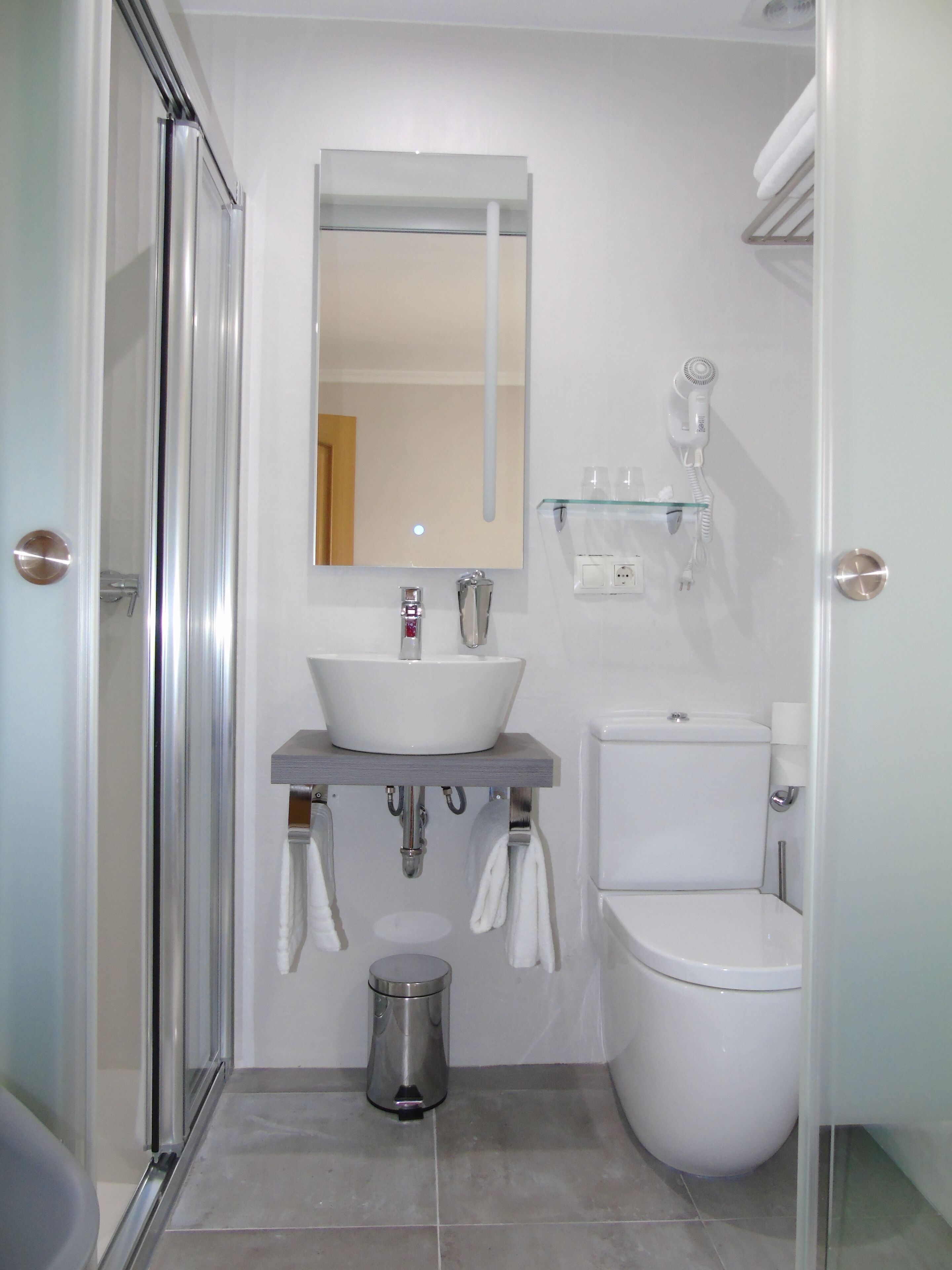A compact bathroom with a glass shower enclosure, a white sink, a toilet, and a small trash bin.