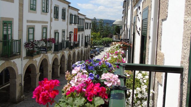 A narrow street with buildings on either side, each adorned with flower boxes. - Casa San Nicolás (Portomarin)