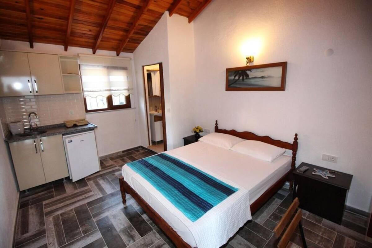 A bedroom with a wooden bed, a nightstand, a small table, and a view of the kitchen area.