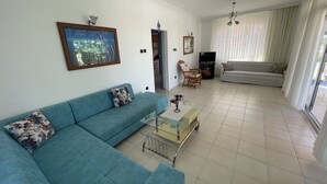 A living room with a blue sofa, a glass coffee table, a wicker chair, a TV, and a large window with curtains. - Kumsal Suit (Marmara)