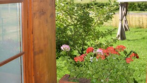 Een houten raamkozijn met uitzicht op een tuin, een bloempot met rode bloemen, en een glimp van een landelijk landschap met een hooibaal en een tent in de verte.