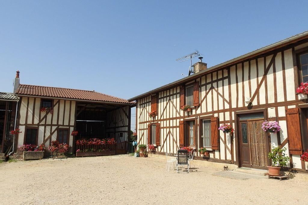 A traditional half-timbered building with a dirt ground in front, a covered area to the left, and potted plants and flowers around.