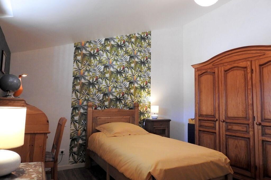 A bedroom with a bed, a wooden wardrobe, a nightstand, a lamp, and a wall with a tropical leaf pattern.