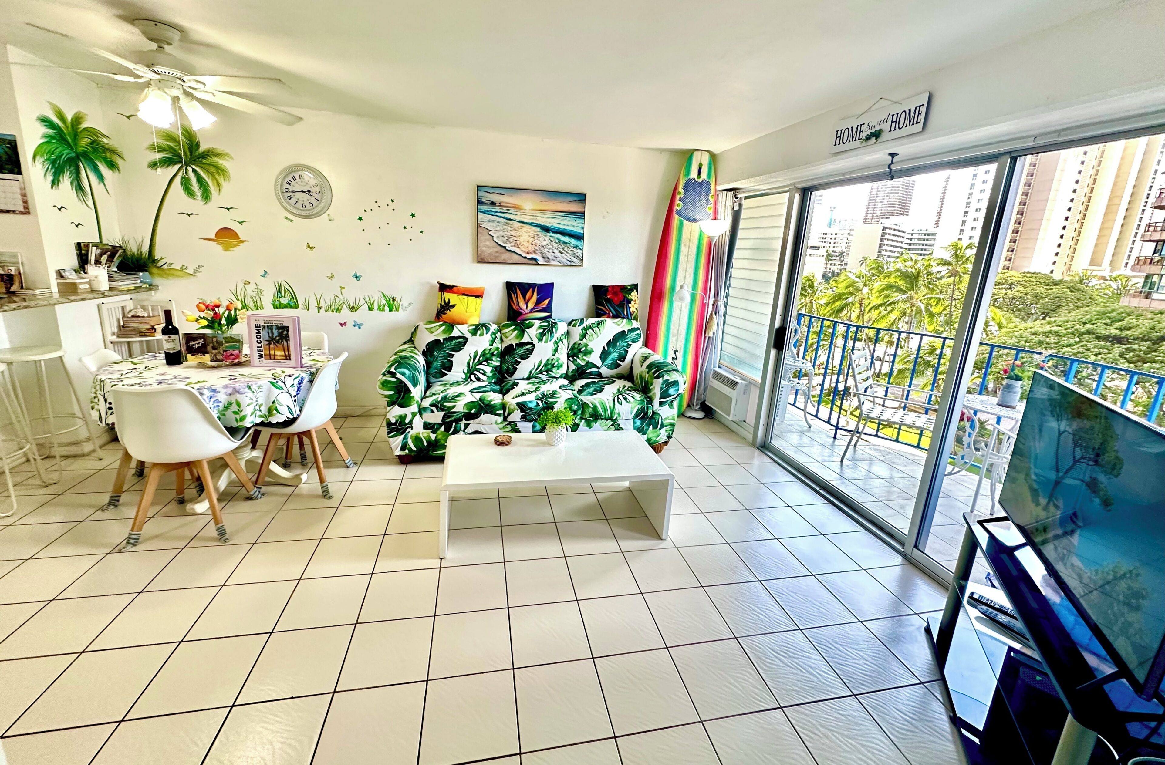 A living room with a sofa, coffee table, dining table, chairs, a TV, and a balcony with a view of buildings and greenery.