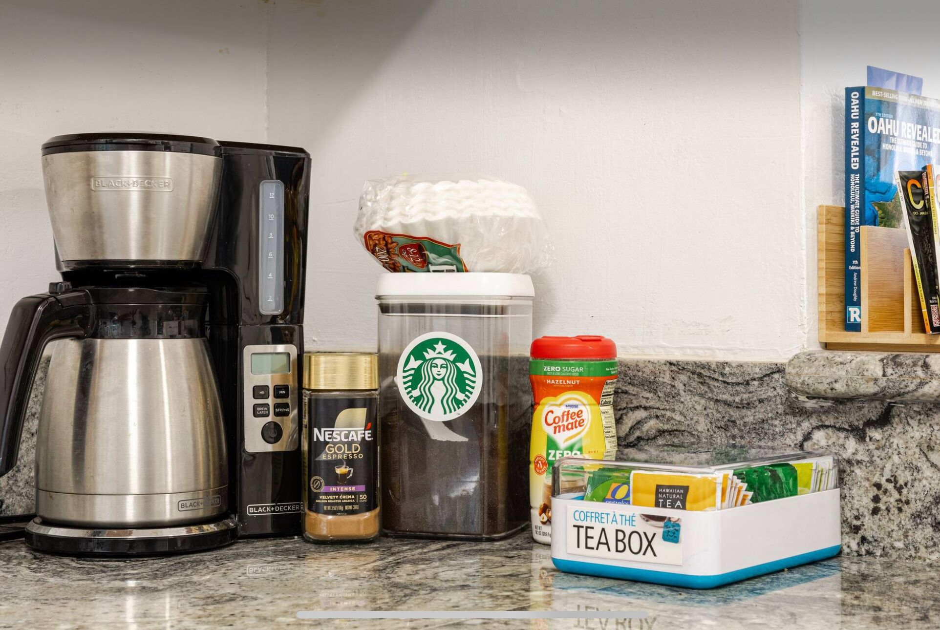 Eine Kaffeemaschine, eine Kaffeemühle, ein Kaffeekocher, ein Starbucks-Kaffeebehälter, ein Nescafe Gold Espresso-Behälter, ein Haselnuss Kaffee-Behälter und eine Tee-Box auf einer Küchenarbeitsplatte.