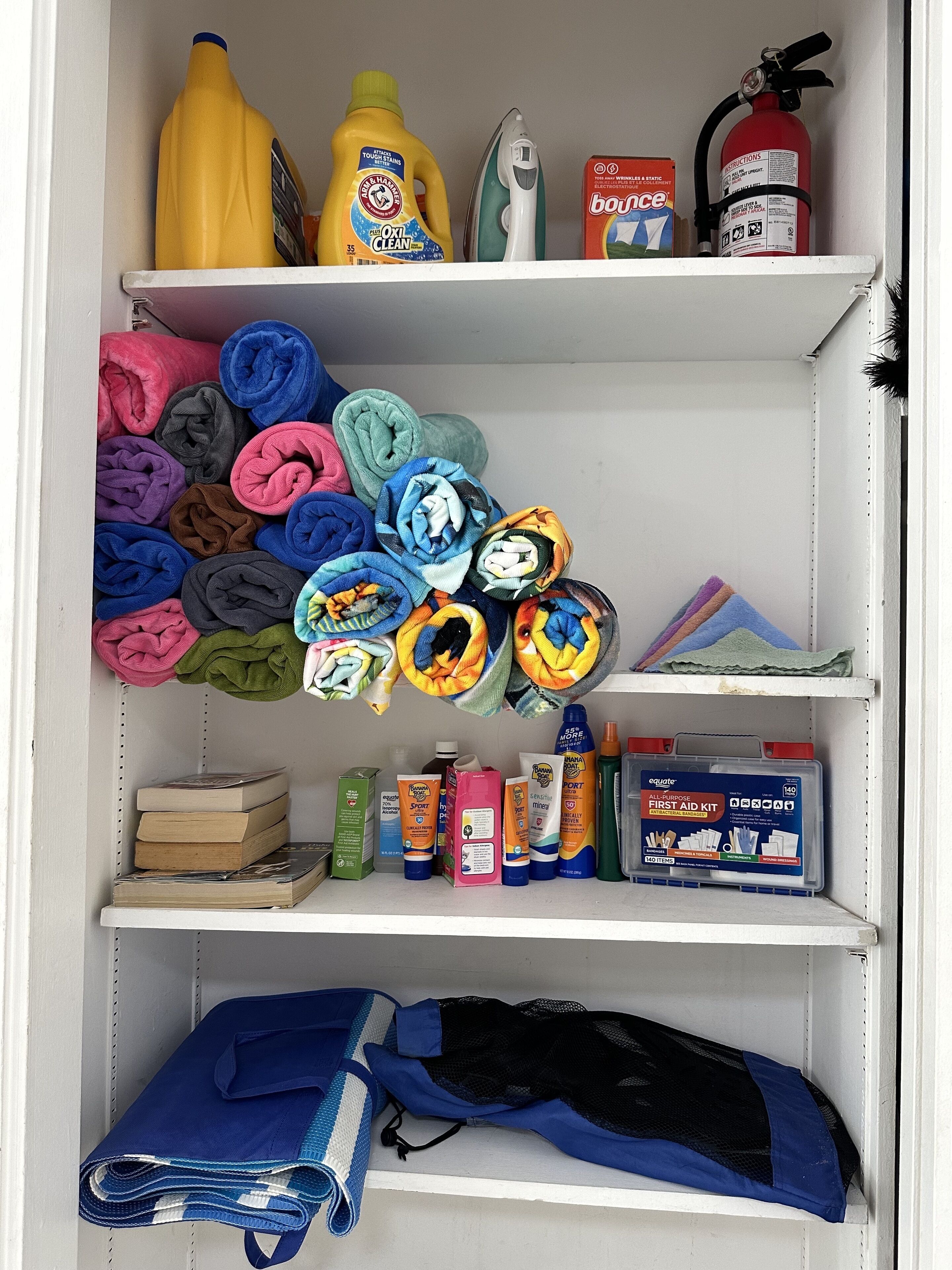 A storage unit with shelves containing folded towels, cleaning supplies, a fire extinguisher, and a first aid kit.